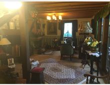 Gemütliches Wohnzimmer im rustikalen Stil mit sichtbaren Holzbalken an der Decke, einem Kamin, antiken Möbeln und dekorativen Elementen wie Teppichen, Vorhängen und Blumen. Ein Fenster mit hellem Vorhang sorgt für natürliches Licht, während eine warme Beleuchtung den Raum erhellt.