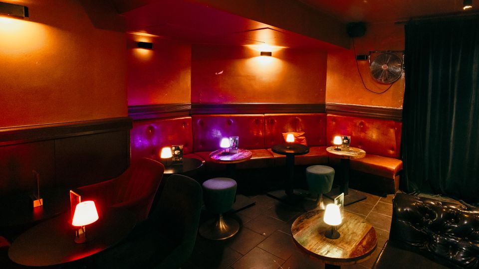 Innenraum einer Lounge- oder Bar-Sitzecke mit gepolsterter roter Rundbank, kleinen runden Tischen und gedimmten Tischlampen. Orangefarbene Wände mit Wandleuchten, dunkler Fliesenboden und ein schwarzer Vorhang an der rechten Seite prägen den Raum.