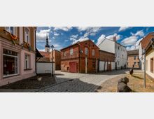 Eine kopfsteingepflasterte Straße in einer Altstadt, umgeben von historischen Gebäuden aus Backstein und verputztem Mauerwerk. Im Hintergrund ist ein Kirchturm mit schwarzer Spitze und vergoldetem Kreuz sichtbar, während bunte Dekorationen und vereinzelte Holzelemente die Umgebung schmücken.