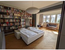 Helles Schlafzimmer mit Parkettboden, Doppelbett und einer wandfüllenden Bücherregalwand. Große Fensterfront mit Sitzbank/kleinem Sofa und Schreibtischbereich sowie einer großen Deckenleuchte.