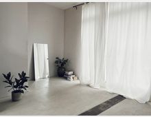 Minimalistisch eingerichteter Raum mit hellem, glattem Boden, großen weißen Vorhängen an bodentiefen Fenstern, einem schmalen Standspiegel an der Wand, einer Zimmerpflanze in einem Topf und einem Bücherstapel neben einer Vase.