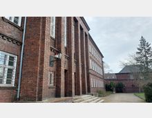 Backsteinbau mit mehreren schmalen, vertikalen Fensterreihen und dekorativen Ziegelmustern an der Fassade. Im Vordergrund Treppenstufen, auf der rechten Seite ein gepflasterter Weg mit angrenzender Grünfläche und Bäumen.