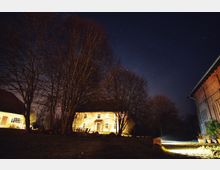 Beleuchtetes Landhaus bei Nacht, umgeben von kahlen Bäumen im Vordergrund und einem sternenklaren Himmel. Rechts sind Teile eines roten Ziegelgebäudes zu sehen, während die Szenerie von Bodenleuchten illuminiert ist.