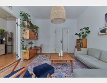 Wohnzimmer mit skandinavischem Einrichtungsstil, gekennzeichnet durch helle Wände, einen Holzfußboden und Möbel aus Holz. Zu den Elementen gehören ein graues Sofa, ein Teppich mit buntem Muster, ein zentraler Couchtisch, Regale mit Büchern und Pflanzen sowie ein geflochtener Lampenschirm.