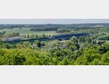 Weitläufige Landschaft mit grünen Feldern, Wäldern und vereinzelten Gebäuden, im Vordergrund dichte Vegetation. Im Zentrum eine blaue Eisenbahnbrücke, die über einen Fluss führt, dahinter Windräder am Horizont.