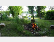 Lagerfeuer auf einer Wiese inmitten von Bäumen und dichtem Grün, mit einem Blick auf einen See im Hintergrund. Im Vordergrund brennt ein Feuer in einer Feuerstelle aus Steinen, und dahinter stehen weiße Gartenmöbel.