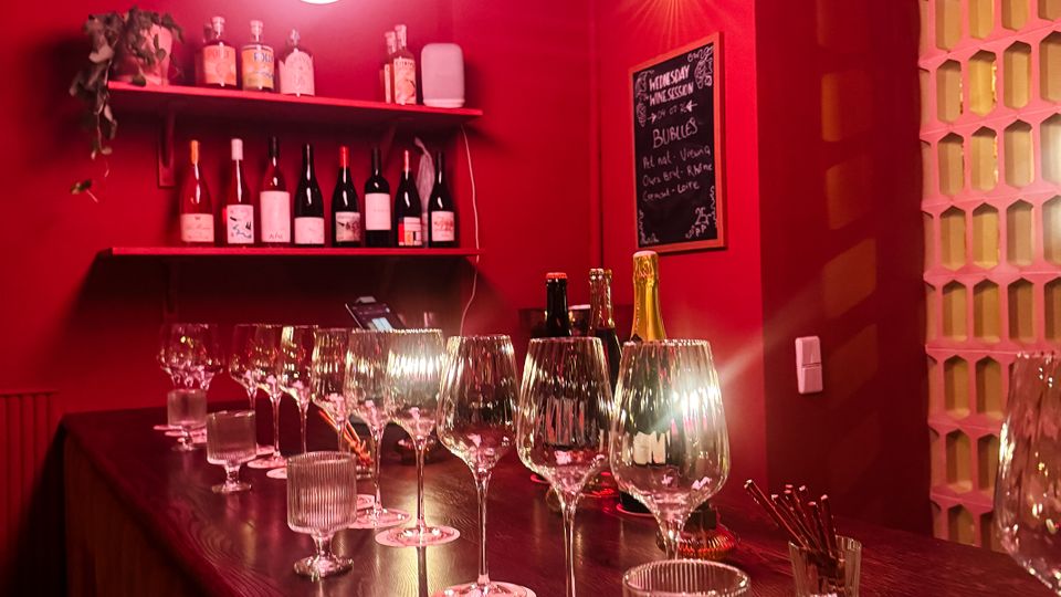 Small bar interior with deep red walls, a long dark wooden counter set with wine glasses and candle holders, and shelves of wine bottles and spirits behind the bar. Round pendant lights hang overhead, and a decorative block-pattern partition lines one side.