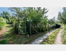 Ein schmaler, unbefestigter Weg führt durch eine grüne Landschaft mit Bäumen und Büschen. Auf der linken Seite befindet sich ein Drahtzaun mit einem offenen Tor, daneben ein Schild mit der Aufschrift "Talweg".