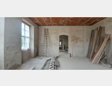 Ein unfertiger Raum mit verputzten Wänden, einer offenen Türöffnung in der Wandmitte und einem rohen Ziegeldach. Links steht ein Fenster mit Plastikfolie, rechts liegen Bretter und Baumaterialien an der Wand, während in der Raummitte eine Leiter und weitere Baustellenutensilien zu sehen sind.