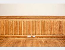 Innenansicht eines Raums mit hoher Holzvertäfelung aus senkrechten Paneelen und breiter Sockelleiste, darüber eine glatte weiße Wand. Vor der Wand liegt ein Holzdielenboden; in der Mitte sind mehrere Steckdosen in die Vertäfelung eingelassen.