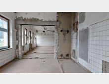 Ein leerstehender, renovierungsbedürftiger Raum mit weißen Wandfliesen, freiliegenden Putzstellen und alten Fenstern. Der Boden zeigt Spuren von Abbrüchen, und an der Decke hängen Kabel und Beleuchtungskörper.