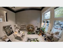 Ein kleiner Laden mit modernem Innendesign, gefüllt mit Pflanzen, Dekorationsartikeln und Büchern. An einer grauen Wand befinden sich Regale mit Keramik, ein runder Holzbogen, und mehrere Fenster bieten Blick auf eine gepflasterte Straße mit geparkten Autos und Bäumen.
