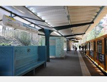 Bahnsteig einer oberirdischen U-Bahn-Station, mit gelbem Zug auf der rechten Seite und überdachter Wartefläche. Links steht eine blaue Sitzbank vor einer Wand mit türkisfarbenen Kacheln, während dahinter Bäume und Sträucher zu sehen sind.