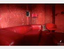 Roter Raum mit rustikalen Ziegelwänden, einer roten Ledercouch, einem roten Stuhl und einer roten Stehlampe. Der Boden und die Wände sind vollständig in Rot gehalten, was dem Raum einen monochromen Charakter verleiht.