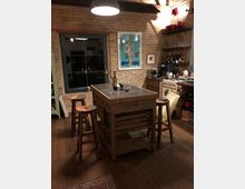 Eine gemütliche Küche mit Backsteinwand, Holzfußboden und einem Holzküchenblock in der Mitte, umgeben von Hockern. An der Wand sind Kunstwerke und Regale mit Gläsern, Gewürzen und Küchenutensilien zu sehen, beleuchtet von einer Deckenlampe.