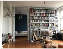 Wohnzimmer mit einer großen Bücherwand aus Metallregalen, gefüllt mit Büchern und Schallplatten. Im Vordergrund stehen ein Sessel, ein kleiner Tisch mit Büchern, und ein Sitzkissen, während ein Kronleuchter und ein Nebenraum mit blau gestrichener Wand sichtbar sind.