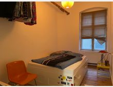 Ein kleines Schlafzimmer mit einem einfachen Bett, das unter der Matratze mehrere Schubladen bietet. An der Wand hängt eine Stange mit Kleidungsstücken, daneben ein orangefarbener Stuhl. Ein Fenster mit halb heruntergelassenem Rollladen und ein gelber Beistelltisch mit Lampe befinden sich im Hintergrund. Holzboden und weiße Wände prägen den Raum.