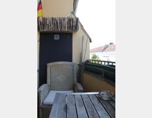 Small apartment balcony with yellow stucco walls, a rustic stick railing above, and a black door panel. A wicker armchair sits beside a weathered wooden table with a glass ashtray, and a railing with planter boxes overlooks nearby buildings.