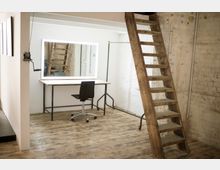 Ein einfach eingerichteter Raum mit rustikalem Holzfußboden, einer Holzwendeltreppe und einer weißen Wand mit einem beleuchteten Spiegel. Vor dem Spiegel stehen ein schlichter Schreibtisch und ein schwarzer Bürostuhl.