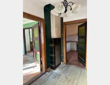 Ein Innenraum mit abgenutztem Holzboden, einer alten, gefliesten Ofenanlage mit grün-schwarzen Kacheln und Holztüren mit eingesetztem strukturiertem Glas. Im Hintergrund sind eine Küche mit weißen Schränken sowie ein weiterer Raum mit grün gestrichenen Wänden sichtbar.