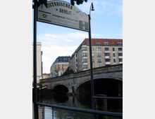 Schild mit der Aufschrift "Historischer Hafen Berlin – Anlegestelle Fischerinsel" im Vordergrund, dahinter eine Steinbrücke über einen Fluss und umliegende Wohngebäude mit roten und schwarzen Dächern.