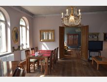 Esszimmer in einem historischen Gebäude mit Holzboden, einem Holztisch mit Stühlen und einem roten Tischläufer. An der Wand hängen vergoldete Spiegel, während durch geöffnete Holztüren ein weiterer Raum mit antikem Schrank und Kronleuchter zu sehen ist.