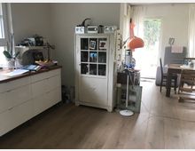 Helles Wohnzimmer mit Holzboden, einer weißen Kommode und einem weißen, verglasten Schrank. Im Raum befinden sich außerdem ein Esstisch mit Stühlen, eine orangefarbene Stehlampe und ein großer Zugang zu einem Garten mit Blick durch eine Glastür.