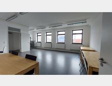 Heller Meetingraum mit mehreren Fenstern, weißen Wänden und grauem Bodenbelag, möbliert mit Holztischen und Stühlen. Im Hintergrund steht ein Regal, und links ist eine offene Tür zu einem angrenzenden Raum zu sehen.