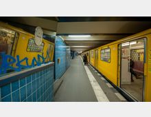 U-Bahn-Station mit gelbem U-Bahn-Zug, der an einem Bahnsteig steht. Die Wände der Station sind mit blauen Fliesen verkleidet, und Beleuchtung hängt an der Decke; im Hintergrund sind wenige Fahrgäste und eine Uhr zu sehen.