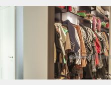 Ein begehbarer Kleiderschrank mit einer Reihe von hängender Kleidung in verschiedenen Farben und Stilen sowie offenen Regalen mit Taschen und Accessoires. Links daneben befindet sich eine geschlossene, weiße Tür.