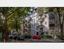 Wohnstraße mit mehrstöckigen Altbauwohnungen, Balkonen und verzierten Fassaden. Im Vordergrund stehen parkende Autos, und große Bäume säumen die Straße, die teils im Schatten liegt.