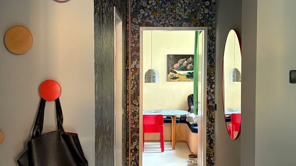 Narrow interior hallway with gray walls, a patterned feature wall around a doorway, and a rug leading into a bright dining area with red chairs. Round wall hooks and a hanging bag are on the left wall, and an oval wall mirror is mounted on the right.