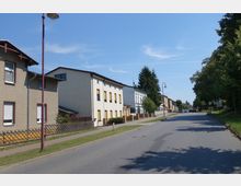 Straßenansicht eines Wohngebiets mit freistehenden Häusern, teils in moderner, teils in traditioneller Bauweise. Links befinden sich Gehwege und Vorgärten mit Zäunen, rechts eine Baumreihe entlang der Straße, die in der Ferne geradeaus führt.