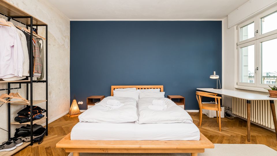 Helles Schlafzimmer mit Doppelbett und Holzrahmen vor einer dunkelblauen Akzentwand, flankiert von zwei Nachttischen. Rechts stehen ein Schreibtisch mit Stuhl vor großen Fenstern und Heizkörper, links ein offenes Kleiderregal; der Boden ist im Fischgrätparkett verlegt.