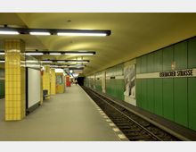 U-Bahnhof mit gelb gekachelten Säulen, einer grünen Wandverkleidung und der Beschilderung "Eisenacher Straße". Der Bahnsteig ist gut ausgeleuchtet, mit Sitzbänken, Werbetafeln und einer Anzeigetafel im Hintergrund.