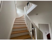 Holztreppe mit weißen Wänden und weißem Geländer in einem hellen Innenraum, die nach oben führt. Im Hintergrund sind Türen sowie ein Fenster mit einfallendem Tageslicht zu erkennen.