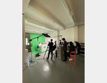 Innenansicht eines Filmstudios mit großer Greenscreen-Wand, Studiobeleuchtung, Kamerastativen und zahlreichen Kabeln. Im Raum stehen außerdem ein weißes Podest vor dem Greenscreen sowie Technik- und Arbeitsbereiche mit Möbeln und Geräten.