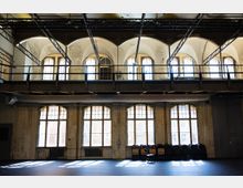 Große, hohe Industriehalle mit zwei Ebenen, hohen Rundbogenfenstern und einer umlaufenden Stahl-Metallgalerie unter der Decke. Sichtbare Trägerkonstruktionen und eine gealterte Putzwand prägen den Raum, am Boden stehen gestapelte Stühle vor den Fenstern.