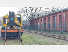 Gelbe Rangierlokomotive, die auf einem Gleis vor einem roten Backsteingebäude mit hohen Fenstern steht. Das Gelände ist von einem Metallzaun umgeben und im Hintergrund stehen kahle Bäume.