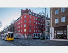 Straßenszene mit einer gelben Straßenbahn auf einem Gleis, einem roten Eckgebäude mit mehreren Stockwerken und Giebeldach sowie umliegenden Gebäuden. Verkehrsschilder, parkende Autos und kahle Bäume betonen die städtische Umgebung.