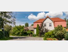 Wohnhaus mit Satteldach und roten Ziegeln, umgeben von dichtem Gartenbewuchs und Bäumen. Im Vordergrund eine ruhige Straße, an der ein Straßenschild und eine Holzbank stehen.