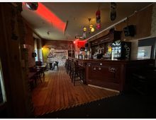 Innenansicht einer schmalen Bar mit langem Holztresen und Barhockern, Holzfußboden sowie kleinen Tischen an der linken Wand. Gedämpftes Licht mit roten Akzenten, Graffiti an den Wänden und Skateboards als Deckendekoration prägen den Raum; hinter dem Tresen stehen Regale mit Flaschen und ein beleuchteter Kühlschrank.