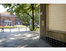 Straßenszene mit einer Kopfsteinpflasterfläche im Vordergrund und einem roten Backsteingebäude mit großen Fenstern und der Aufschrift „Sonnenallee“ im Hintergrund. Ein Baum und parkende Autos befinden sich rechts, Blumenbeete säumen den Platz vor dem Gebäude.