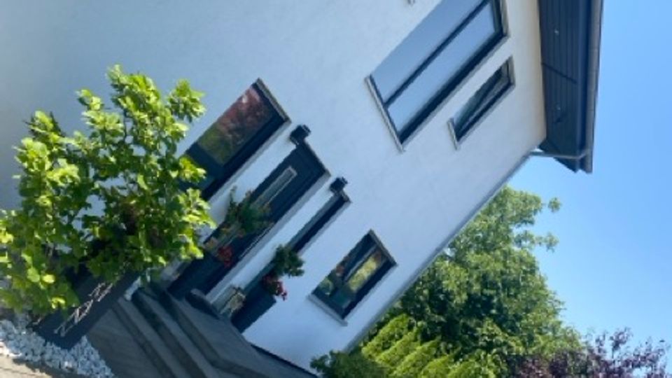 Schräg aufgenommene Außenansicht eines modernen weißen Wohnhauses mit großen rechteckigen Fenstern und dunklem Dachüberstand. Davor liegt eine gepflasterte Einfahrt mit kleinen Büschen und Bäumen sowie angrenzendem Grünbereich.