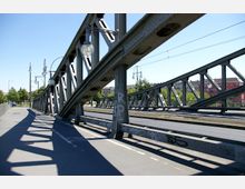 Stählerne Fachwerkbrücke mit schrägen Verstrebungen und Straßenlaternen, daneben ein Geh- und Radweg. Im Hintergrund sind Bäume sowie Wohnhäuser und ein rotes Gebäude zu sehen.