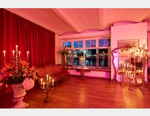 Eleganter Innenraum mit roten Samtvorhängen, antikem Sofa, poliertem Holzboden und zahlreichen Kerzenleuchtern sowie Blumenarrangements. Große Fensterfront mit Blick auf ein beleuchtetes Ufer bei Nacht und ein großer vergoldeter Spiegel an der Wand.