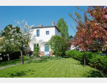 Zweistöckiges Wohnhaus mit weißer Fassade und blauen Türen, umgeben von einem gepflegten Garten mit blühenden Obstbäumen, Sträuchern und einer Rasenfläche unter klarem, blauem Himmel.