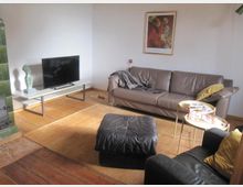 Wohnzimmer mit Holzdielenboden, einem grünen Kachelofen am Rand, einem braunen Ledersofa, einem schwarzen Hocker und einem kleinen weißen TV-Tisch mit Flachbildfernseher. An der Wand hängt ein gerahmtes Kunstbild, und ein kleiner Beistelltisch steht neben dem Sofa.