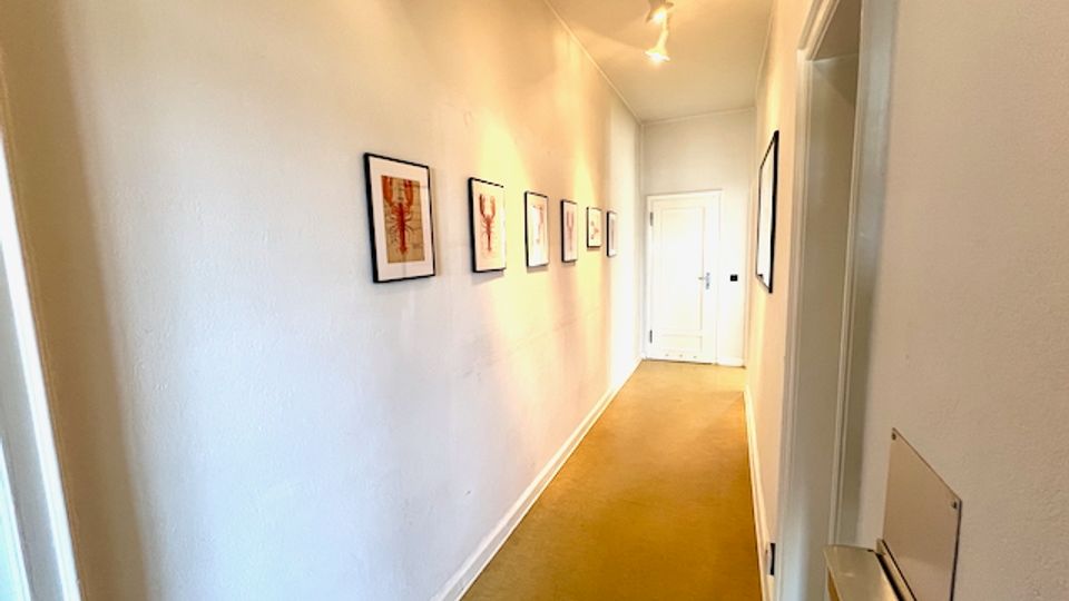 Langer, schmaler Innenflur mit weißen Wänden und beigem Teppichboden, an der linken Wand mehrere gerahmte Bilder. An der Decke sind Spotleuchten montiert, am Ende des Gangs befindet sich eine weiße Tür.