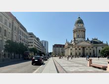 Straßenansicht des Gendarmenmarkts in Berlin mit dem Deutschen Dom rechts im Bild, einem historischen Gebäude mit Säulenfassade und Kuppel. Links ist eine Straße mit parkenden Autos, Bäumen und modernen sowie historischen Gebäuden sichtbar.